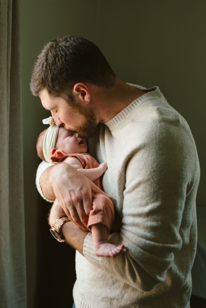 A beareded man holds a newborn baby tenderly and gently kisses its head.